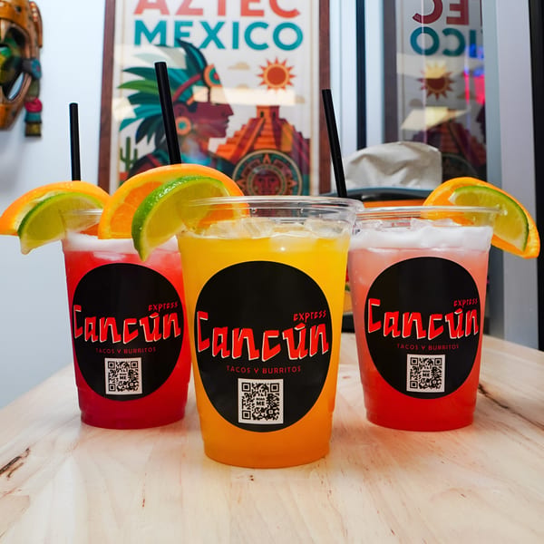 Three vibrant Cancun Express drinks, red and orange, garnished with citrus slices and straws on a wooden table.